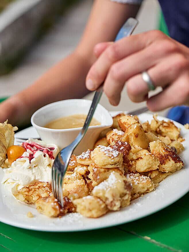 Eine Urlauberin nimmt genüsslich Stückchen vom Kaiserschmarren mit dr Gabel auf. Ein Teller mit Kaiserschmarren