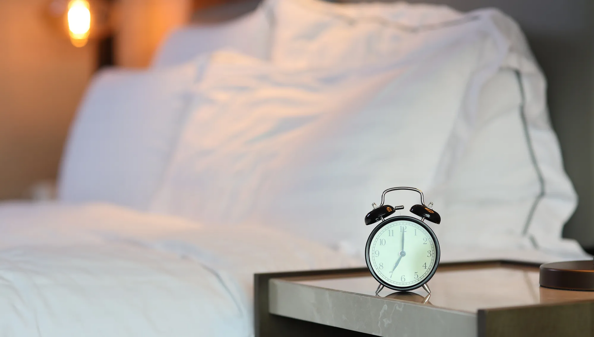 Blick auf ein Bett mit weißen Lacken und einem Wecker auf einem Nachtisch davor Doppelbett frisch bezogen