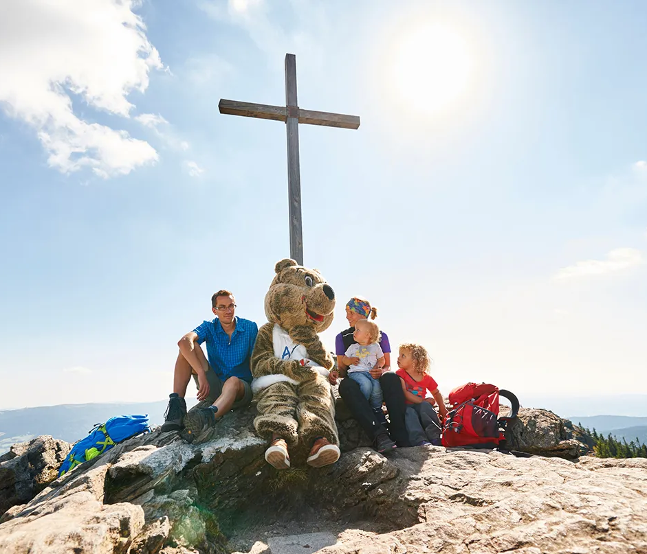 Spaßige Momente am Arbergipfel mit dem Bärenmaskottchen Ein ArBär sitzt mit einer Familie auf einem Felsen, während die Sonne hell am Himmel scheint.